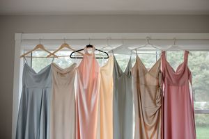 Pastel bridesmaids dresses in blue, blush, and pink tones hang neatly on wooden hangers by a window.