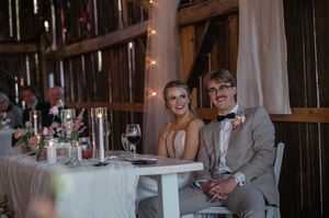 A couple sits together at their rustic barn wedding reception decorated with string lights and floral centerpieces.