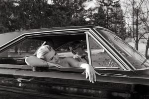 A black and white photo of a relaxed dog looking through the window of a classic car.