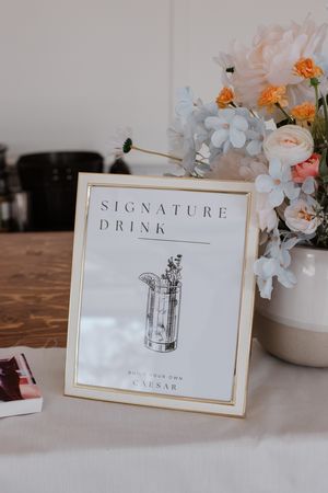 Elegant signature drink menu sign with gold frame sits beside delicate floral arrangement on white table at event.