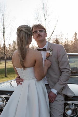A couple in formal wedding attire pose together in front of a vintage car during golden hour.