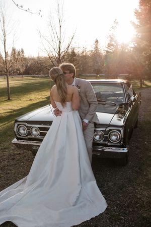 A romantic wedding couple embraces next to a vintage car during golden hour sunset in a scenic outdoor setting.
