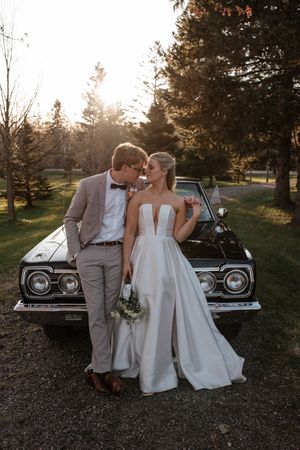 Newlywed couple poses intimately against a classic car during golden hour sunset with pine trees in background.