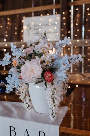 A romantic floral centerpiece in a white vase with pink and white flowers against a backdrop of twinkling string lights.