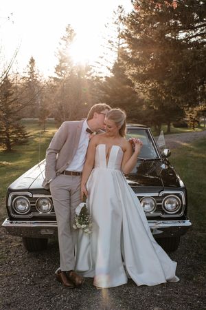 A romantic wedding photo with a couple leaning against a classic black vintage car during golden hour sunset.