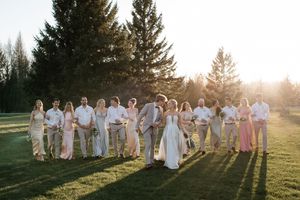 Wedding party gathered outdoors at sunset casting long shadows on grassy field with evergreen trees in background.