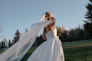 Flowing white wedding dress and veil flutter in the breeze against an evergreen forest backdrop at sunset.