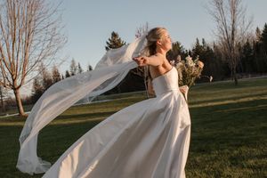 A flowing white wedding dress and veil catch the wind dramatically against an outdoor landscape at sunset.