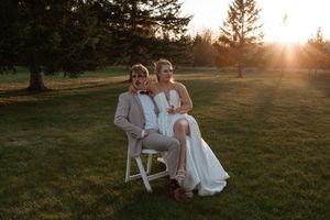 Wedding couple sits together on white wooden chair in sunny garden setting at golden hour.