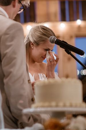 An emotional moment during wedding speeches as someone dabs tears at a reception with a wedding cake visible.