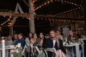 Wedding guests seated at reception tables enjoy entertainment under warm string lights in rustic barn venue.