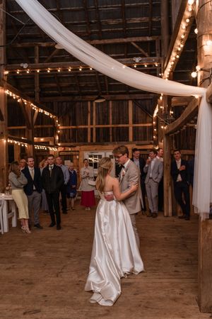 Wedding guests dance together on a rustic barn floor decorated with string lights and white fabric drapes.