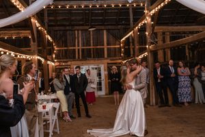Newlyweds dancing with guests in a rustic barn reception venue decorated with string lights.