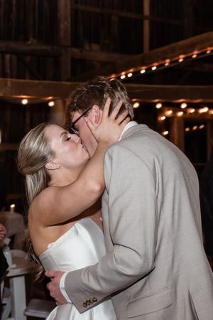 Bride and groom share a romantic kiss during their wedding reception under warm string lights in a rustic venue.