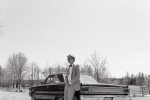 Black and white photo of person standing by vintage car in winter setting.