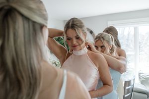 Women in a bridal party help zip up a blush pink halter dress during wedding preparations.