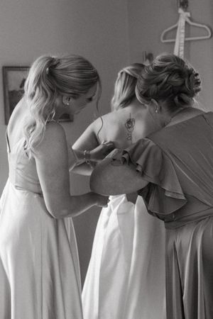 Two people wearing elegant dresses help with wedding preparations in a tender black and white moment.
