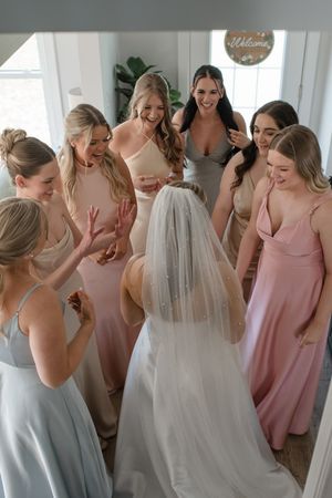 A group of bridesmaids in pastel dresses gather around a bride wearing a white gown and veil in a bright indoor setting.