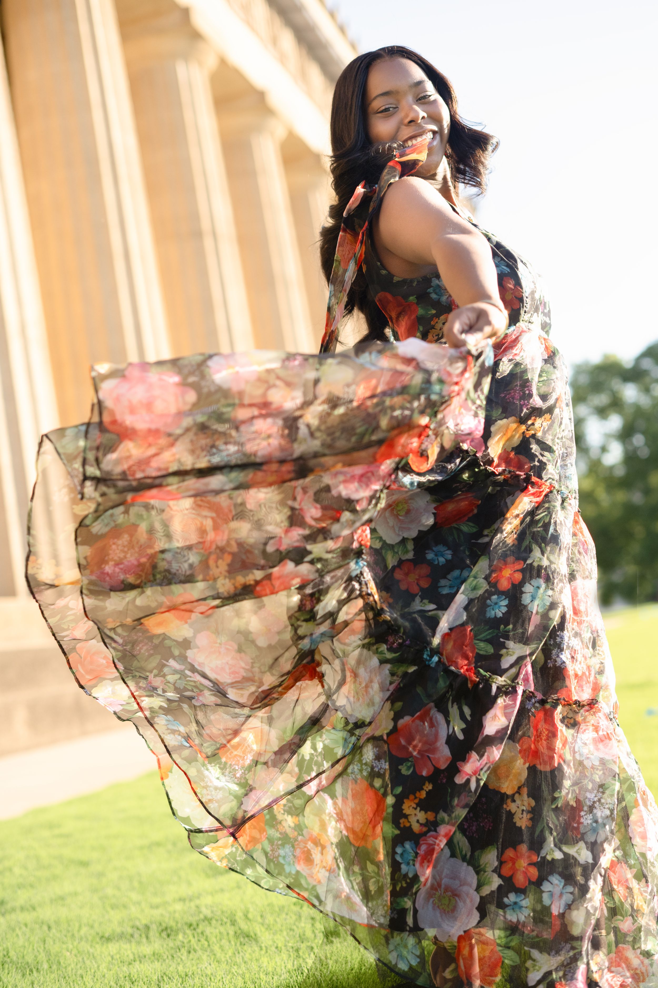 A flowing floral maxi dress twirls gracefully on a sunny day against classic architectural columns and green lawn.