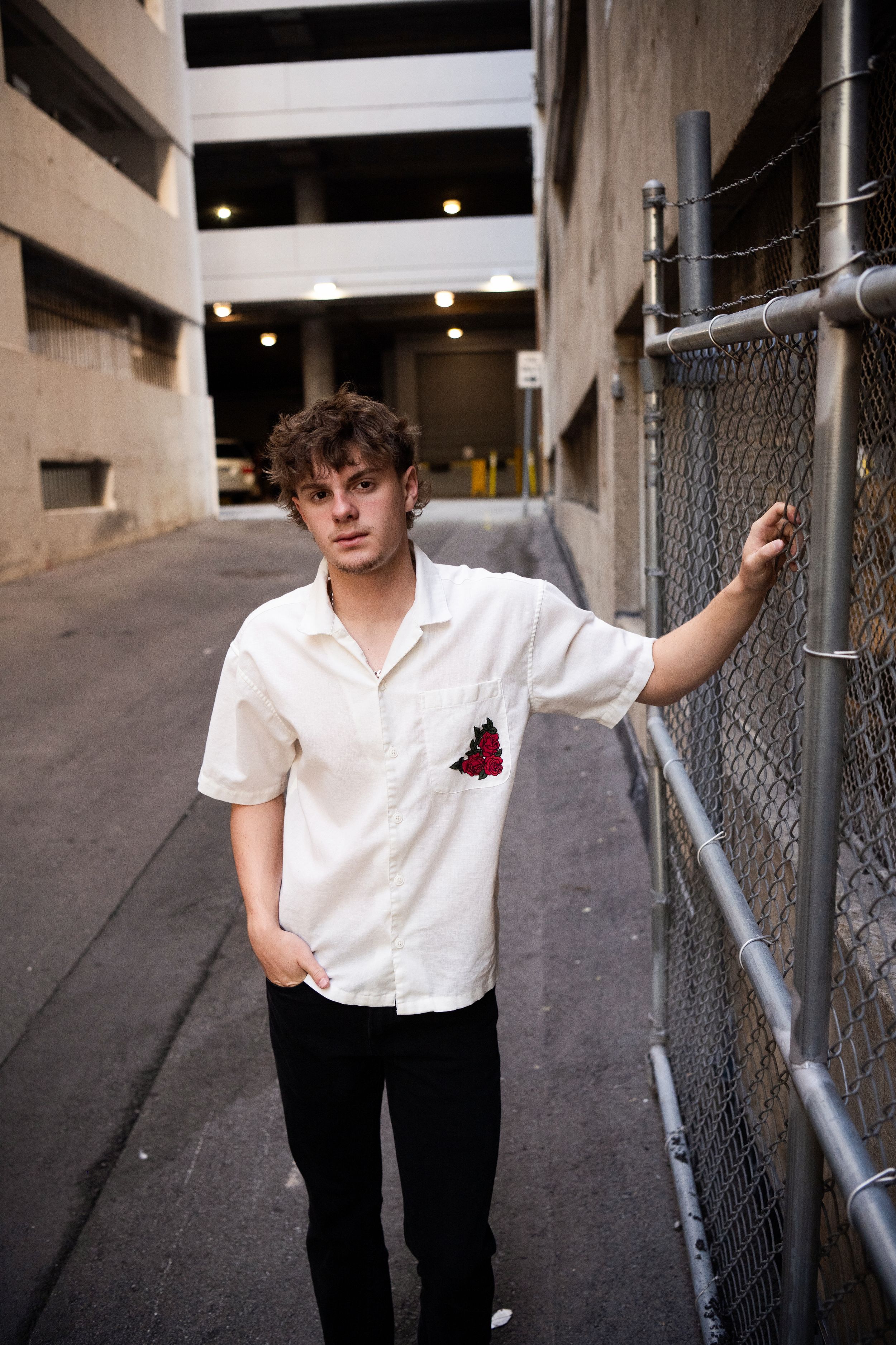 A person in a white button-down shirt with a red detail poses against a chain link fence in a parking garage.