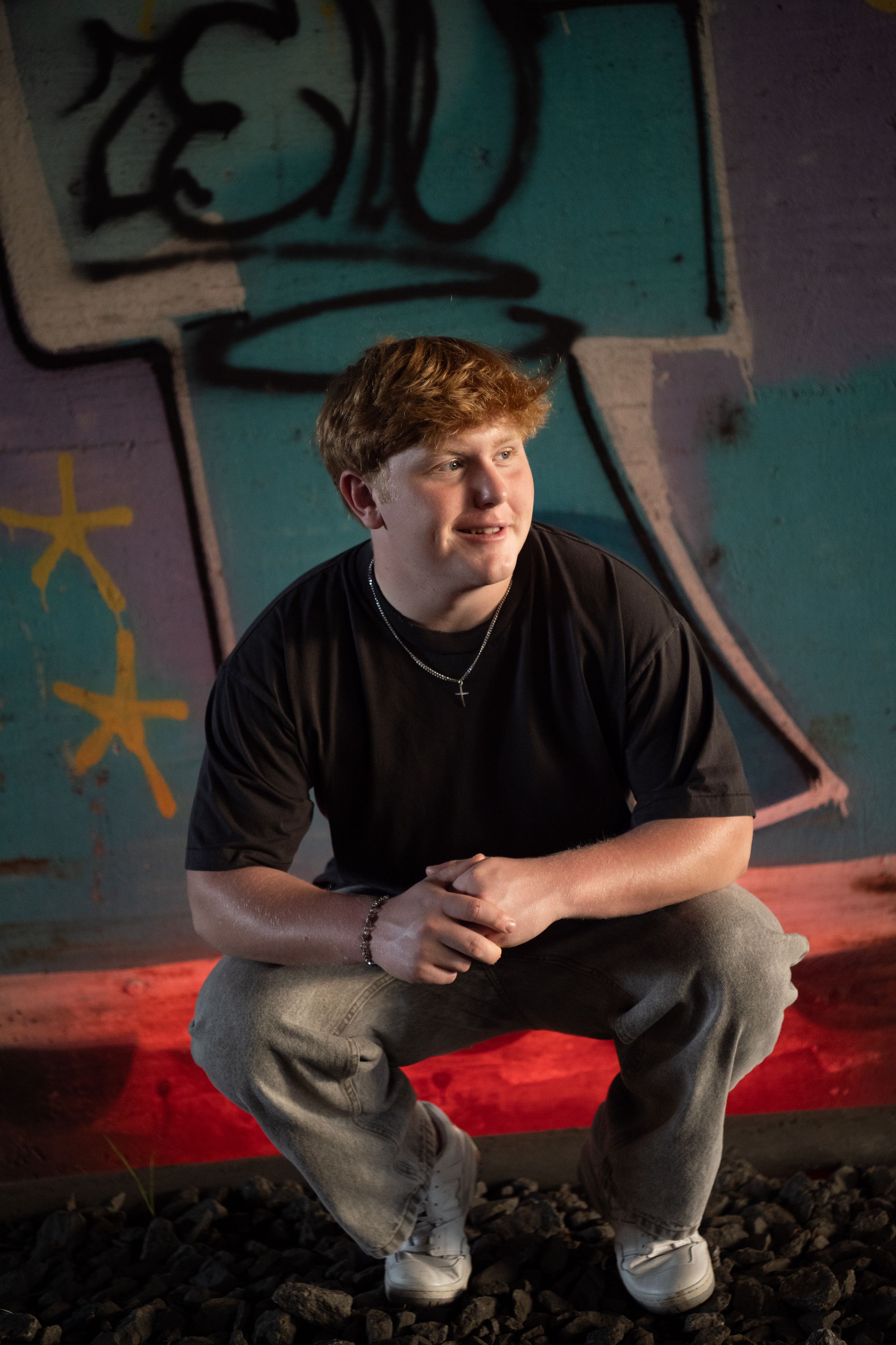 A person in a black t-shirt crouches against a graffiti wall with teal background and red lighting effects.