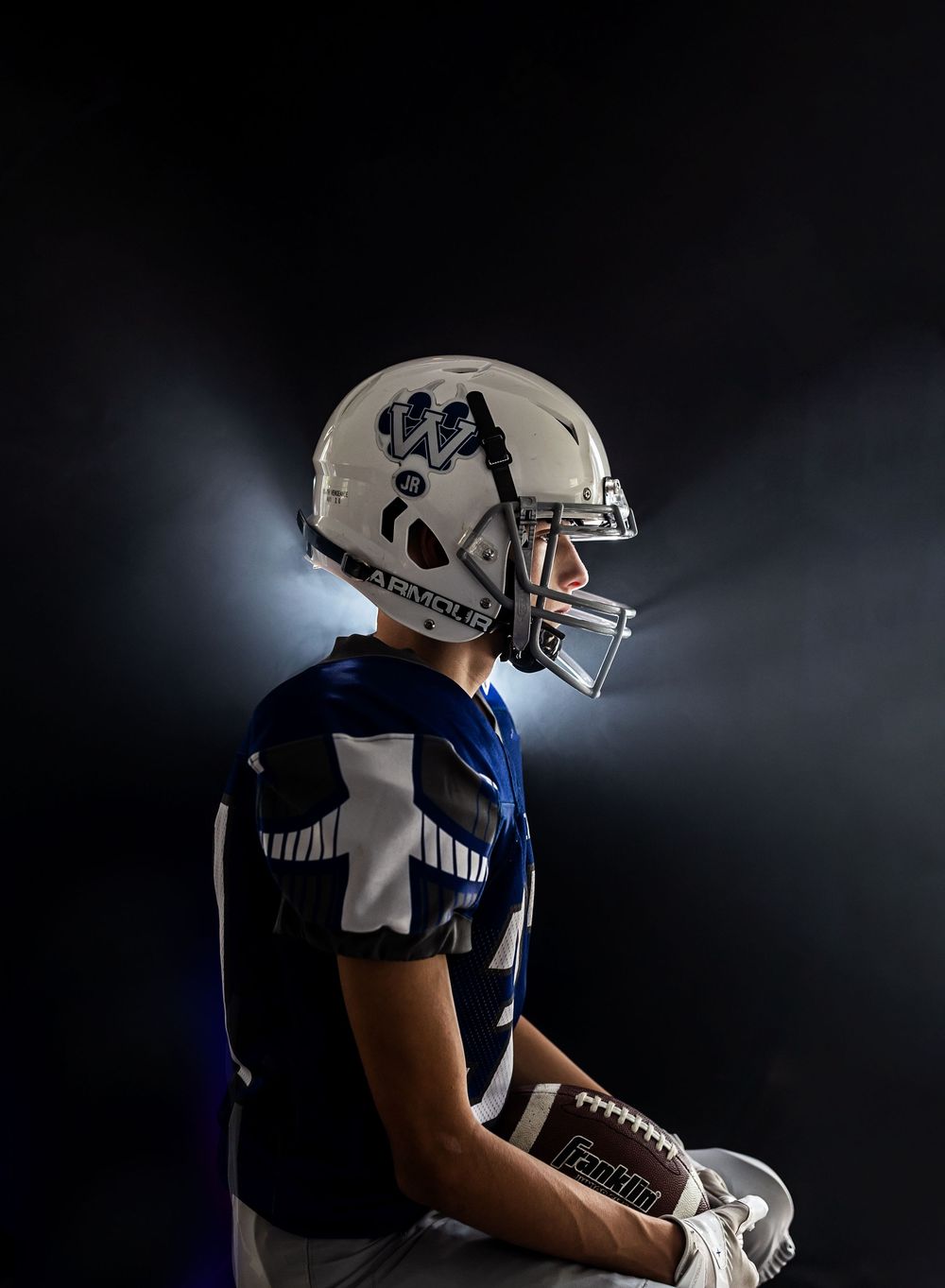 Football player in white and blue uniform stands in profile view against dark background.