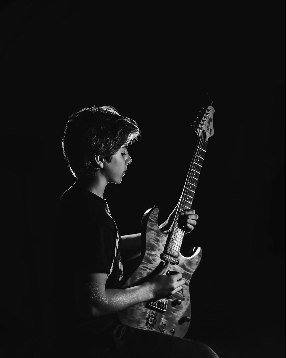 A musician plays an electric guitar in dramatic black and white lighting against a dark background.