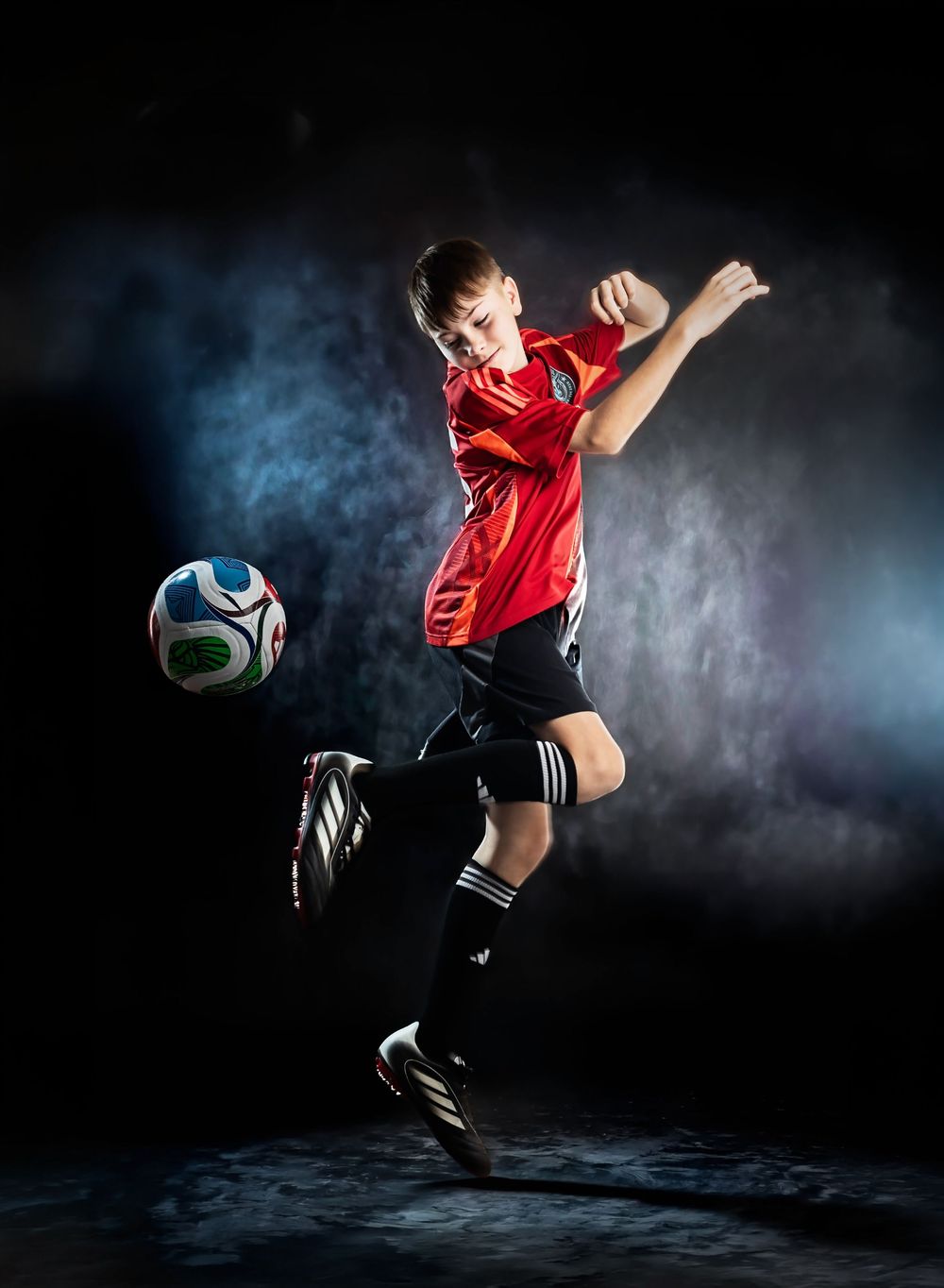 A soccer player in red uniform performs a dramatic kicking motion against a dark background with atmospheric smoke effects.