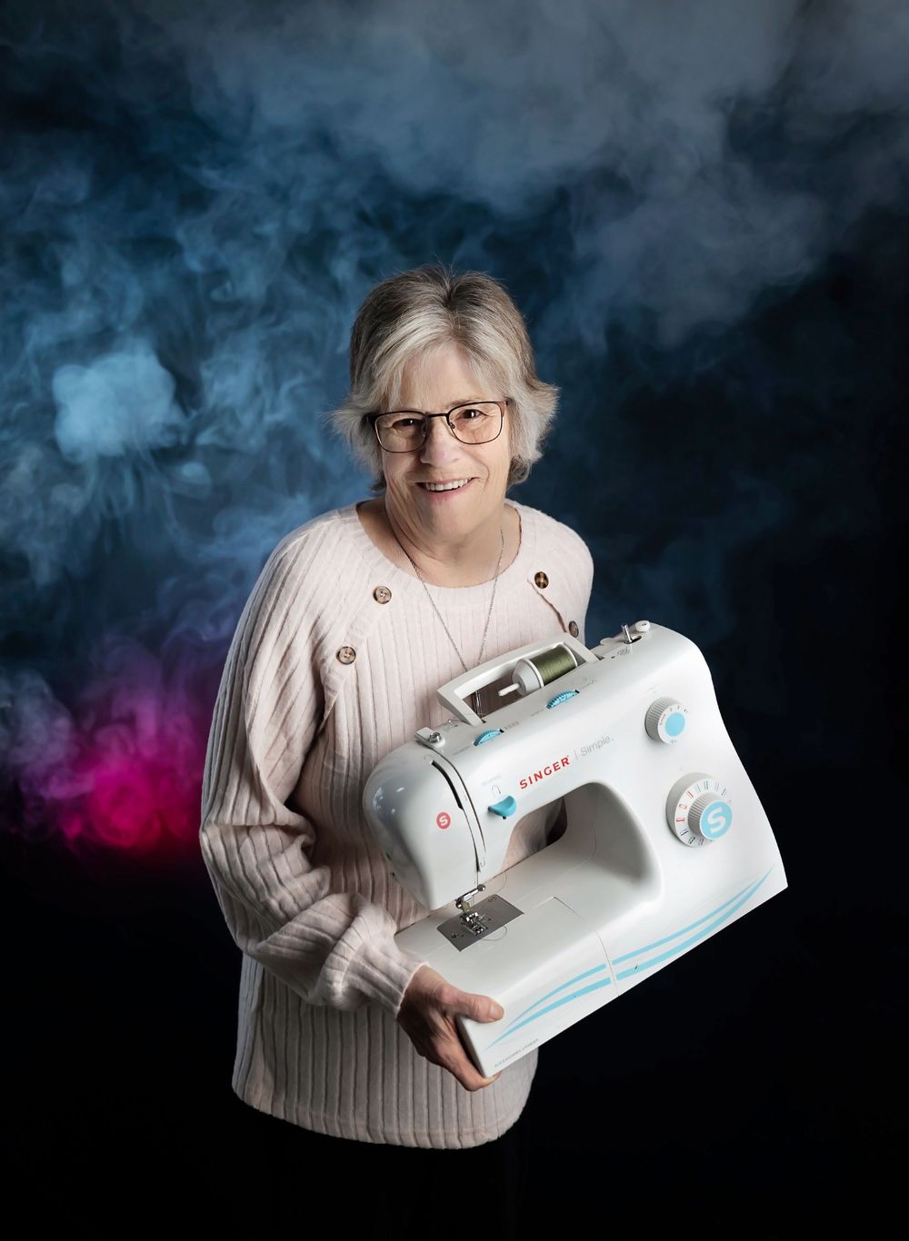 A person in a cream sweater poses with a white sewing machine against a moody dark backdrop with blue and pink smoke.
