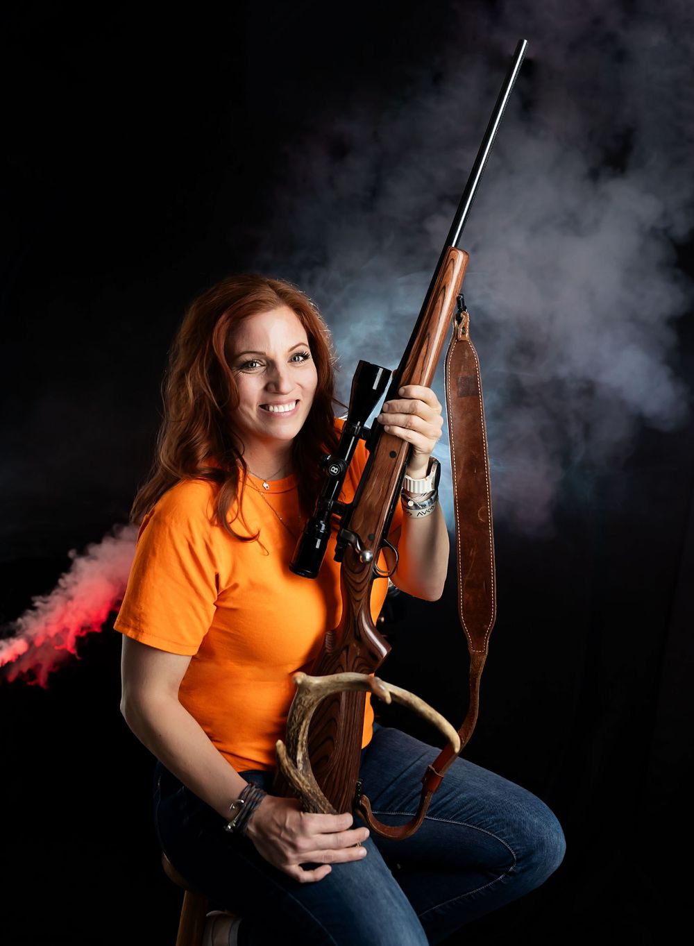 Hunter in bright orange shirt poses with rifle and deer antlers against dark smoky background.