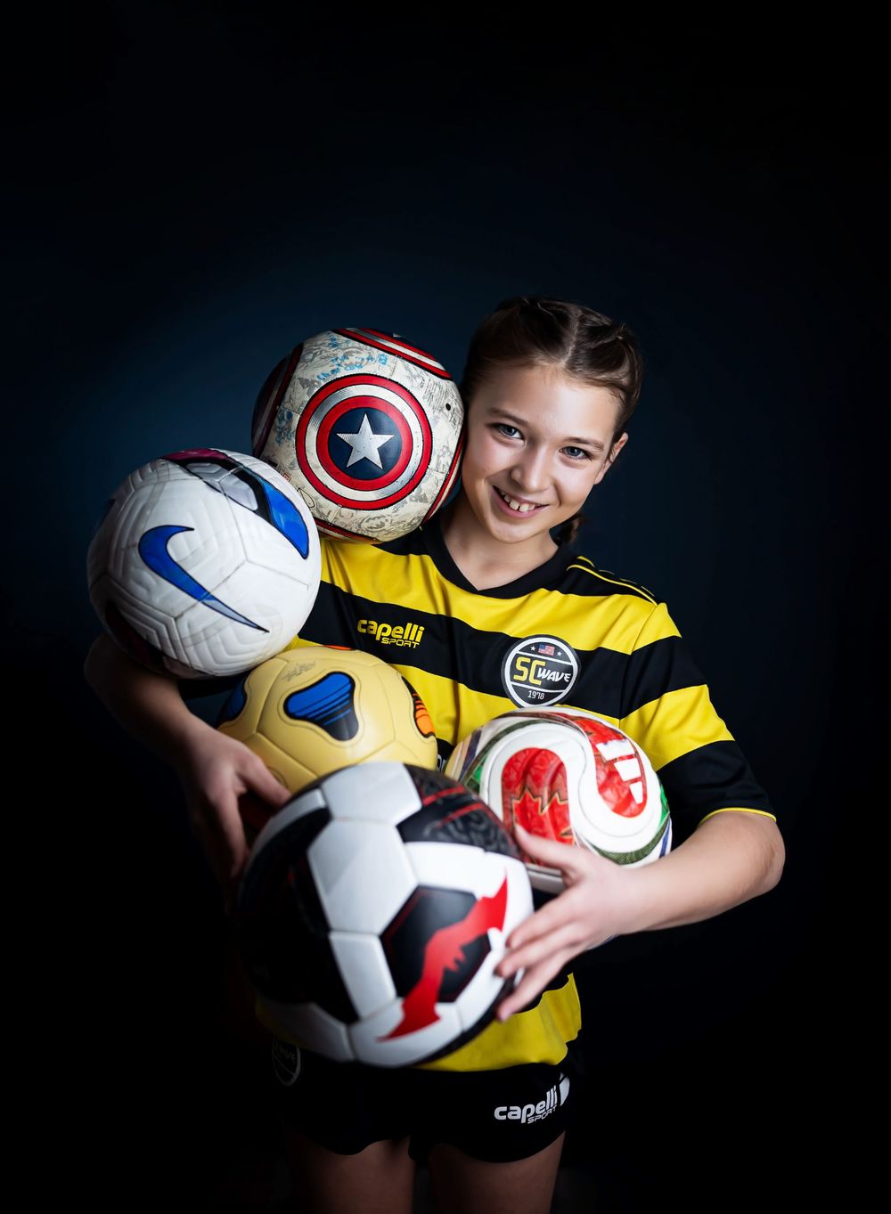 A young athlete in yellow jersey poses with multiple soccer balls against a dark background in a creative sports portrait series.