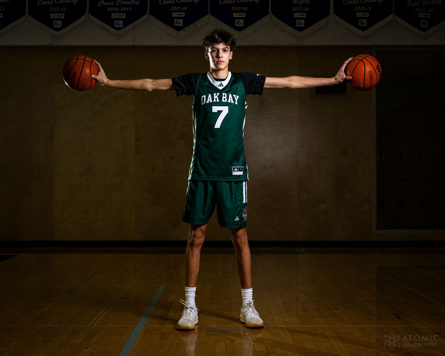 A dramatic photo of a teenage male basketball player posing with basketballs held on outstretched arms.