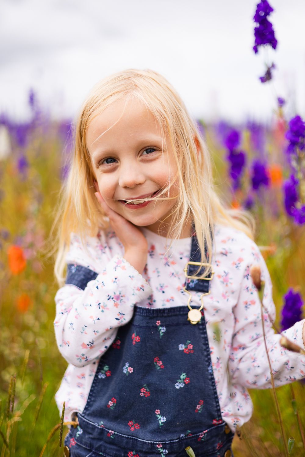 Portrait photo of a young girl amongst purple and red flowers. Her hand on her cheek.
