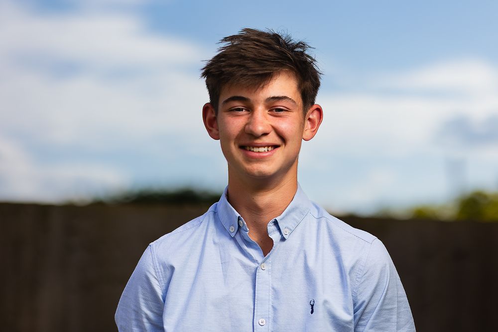 Portrait photo of a teenage boy, smiling and wearing a light blue shirt. Outdoors with a blue sky background.