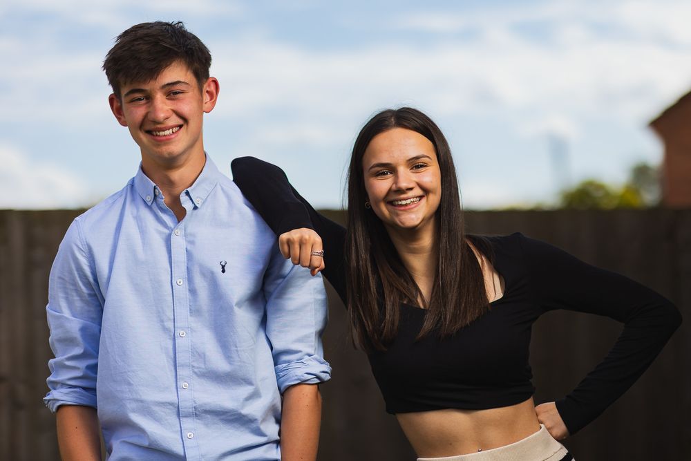 A family portrait photo of a teenage brother and sister. Both smiling and relaxed, wearing smart summer wear outdoors.