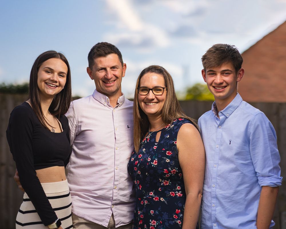 A family portrait photo of a mother, father, son and daughter. All smiling and relaxed wearing smart casual summer wear. Outdoors with a blu