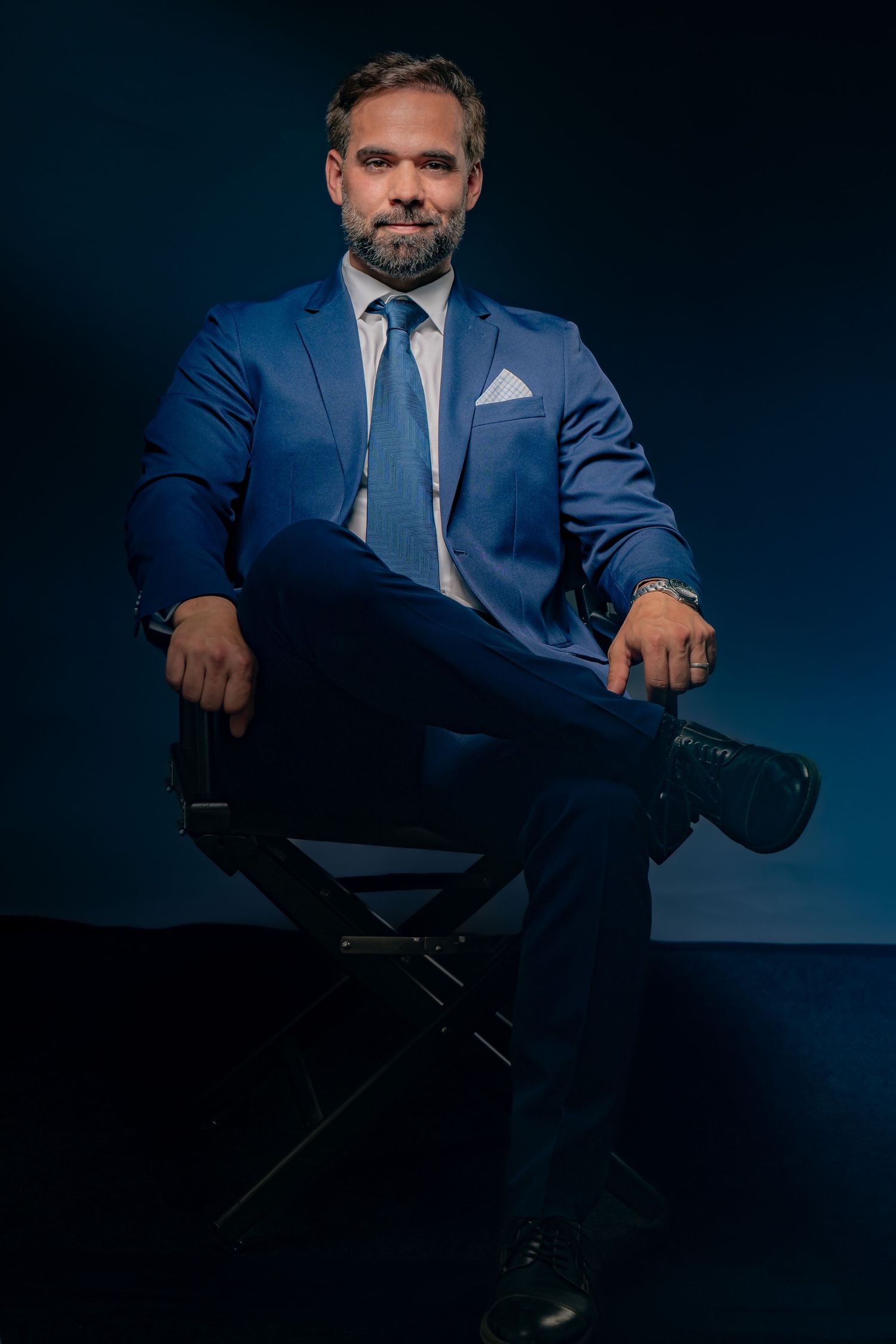 Professional businessman in a tailored blue suit sits confidently against a dark studio backdrop.