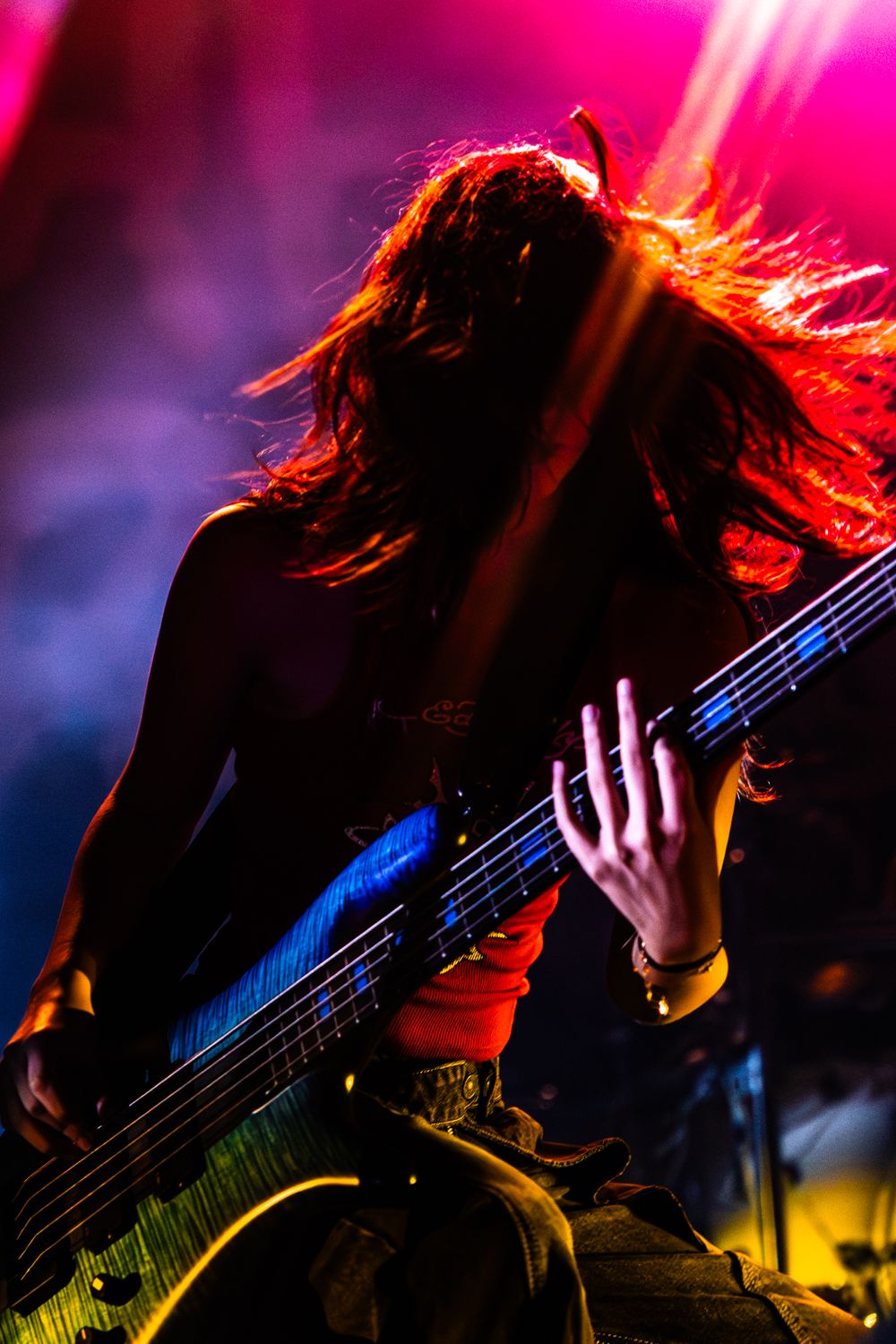 Dynamic concert photo of a bassist performing with flowing hair under vibrant red and purple stage lights.