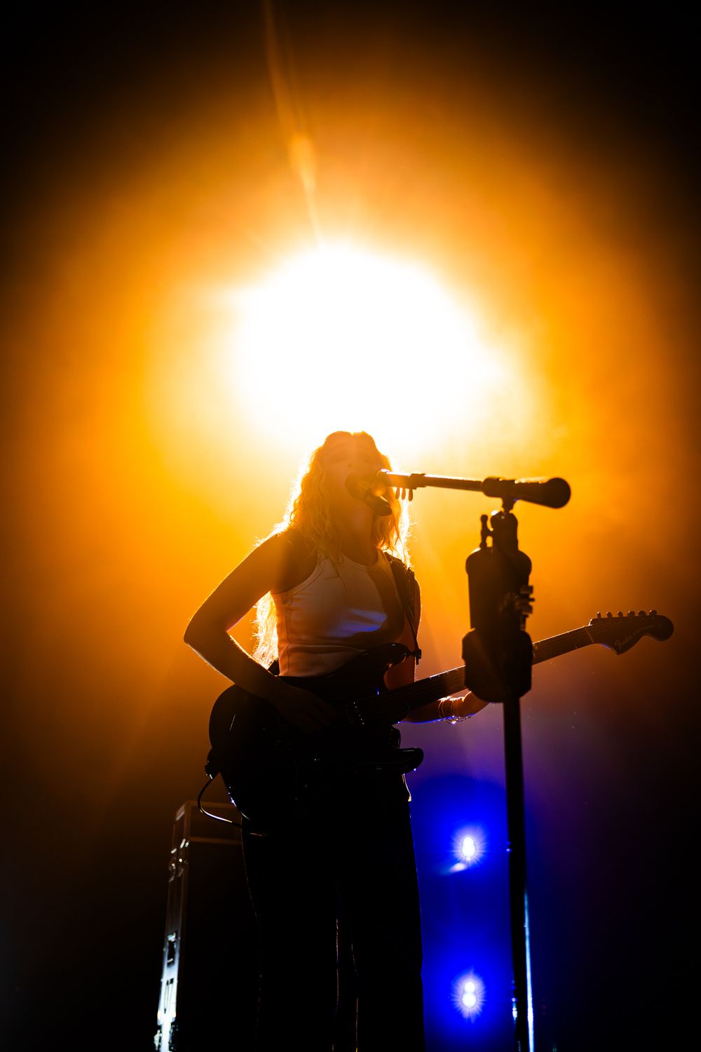 Silhouetted musicians perform on stage against a dramatic orange sunset backdrop with blue accent lighting.