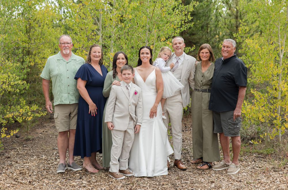 Large wedding party group photo with green foliage and natural scenery in background.