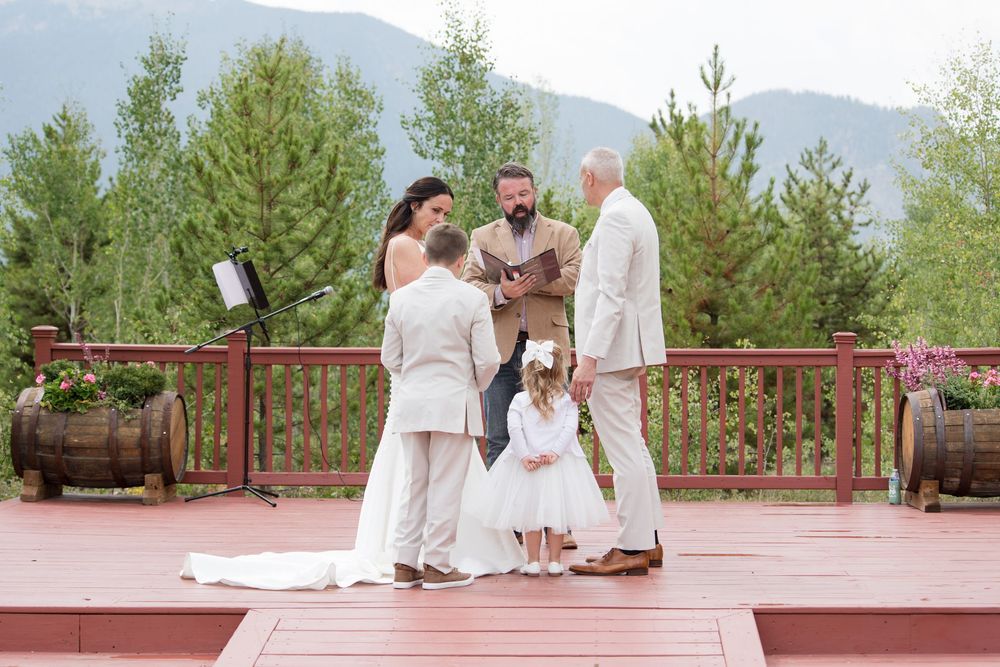 A small group gathers for an intimate outdoor ceremony on a wooden deck surrounded by evergreen trees and mountains.