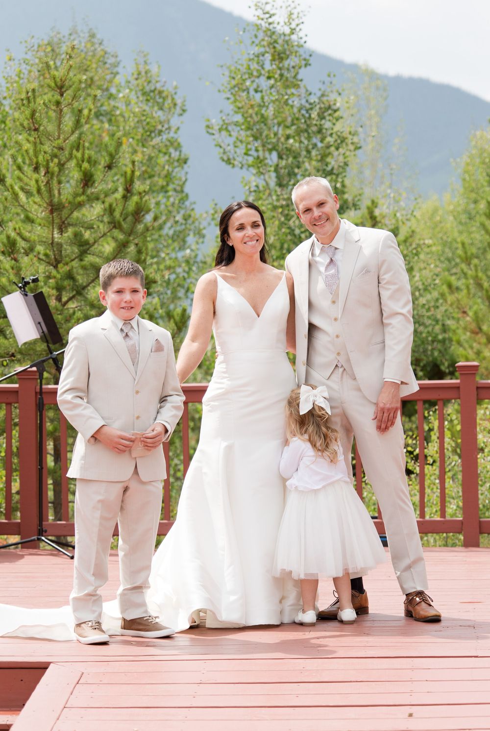 Wedding party in light beige attire poses on wooden deck with mountain backdrop and trees.