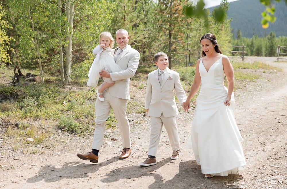 A wedding couple walks along a mountain path surrounded by evergreen trees and natural scenery.