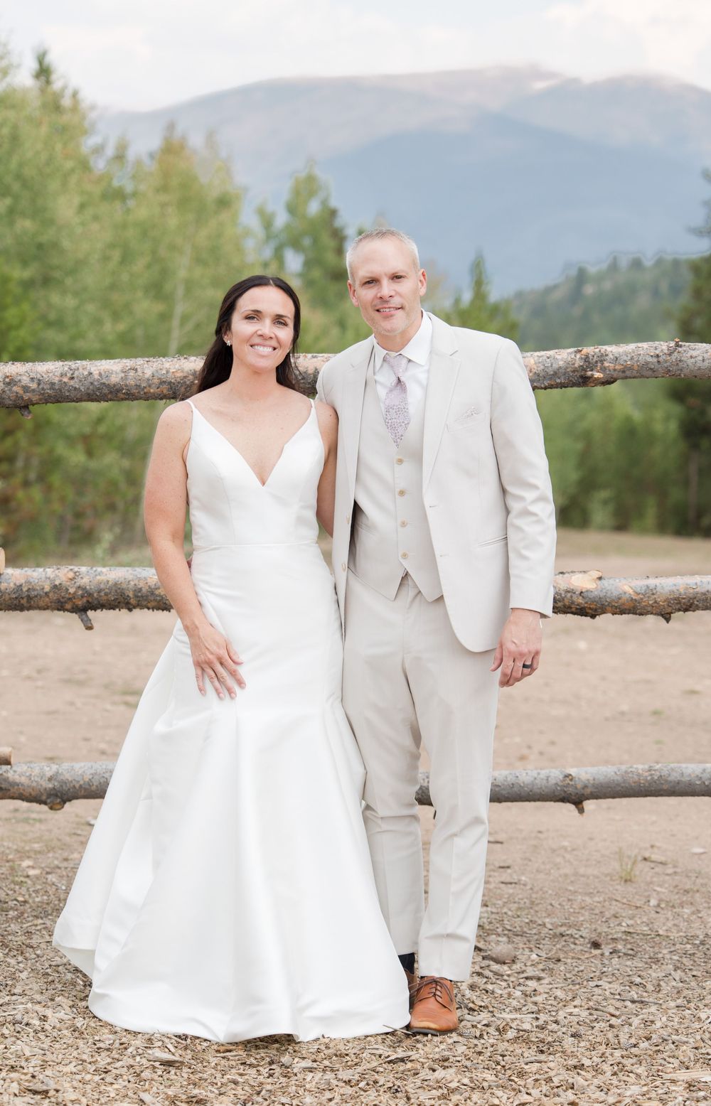 A wedding couple poses in front of a rustic wooden fence with mountain views in the background.
