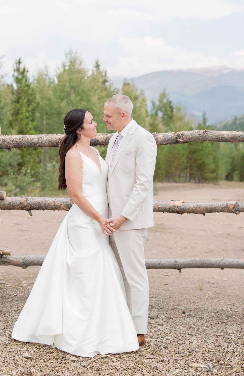 A romantic wedding portrait against a rustic log fence with mountain scenery and evergreen trees.