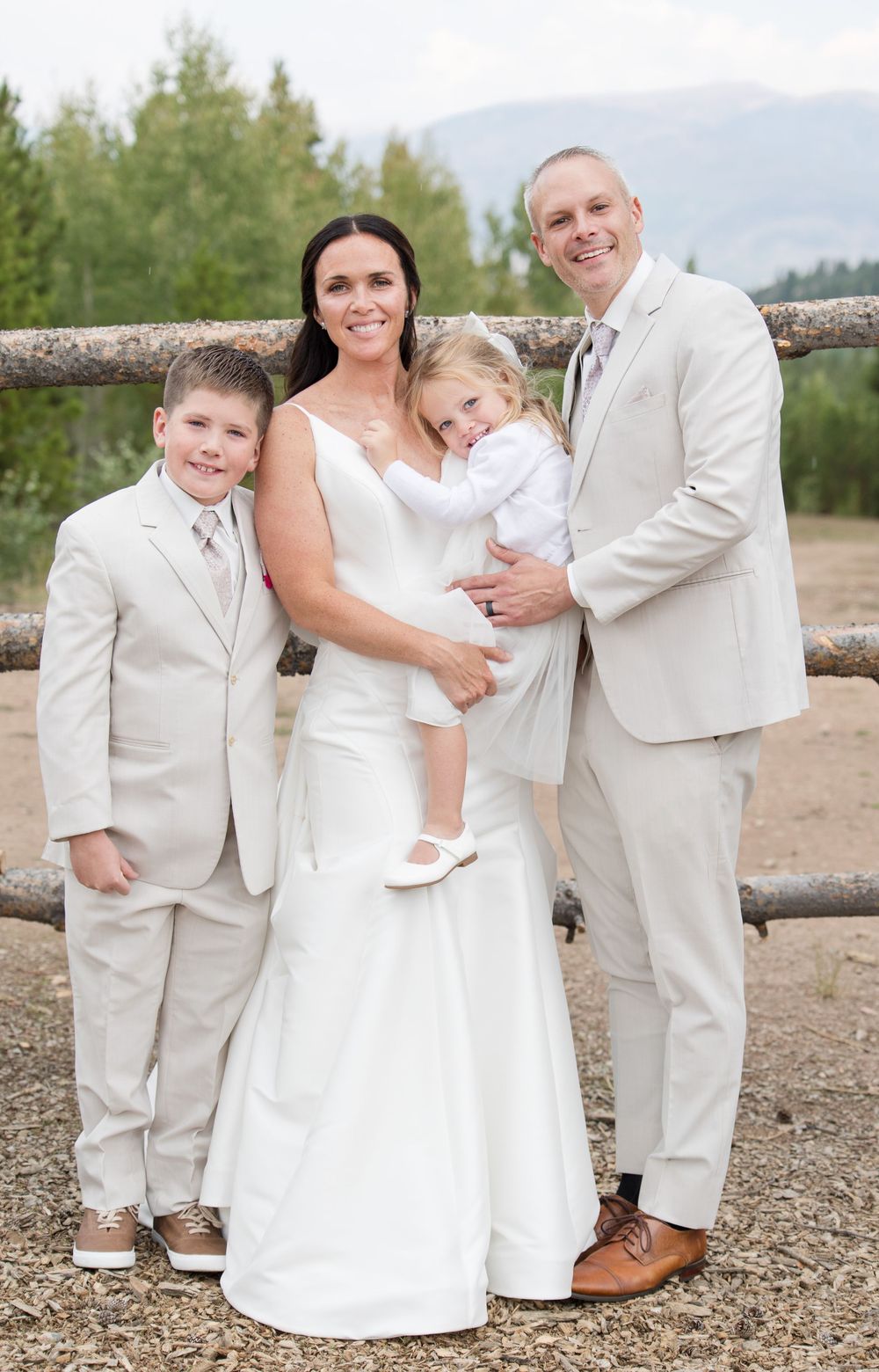 Wedding party poses together by wooden fence with mountains and trees creating scenic backdrop.