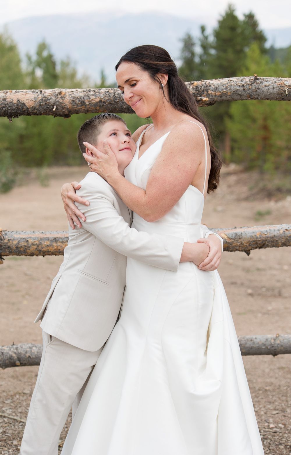 Wedding couple embraces tenderly in front of rustic wooden fence with evergreen trees in background.