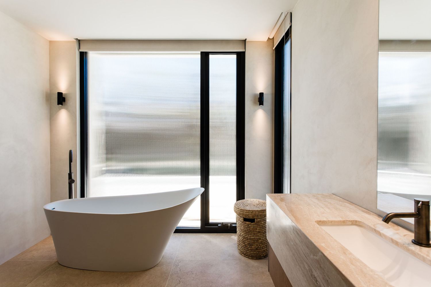 Modern white freestanding bathtub sits beneath large windows in a minimalist bathroom interior.