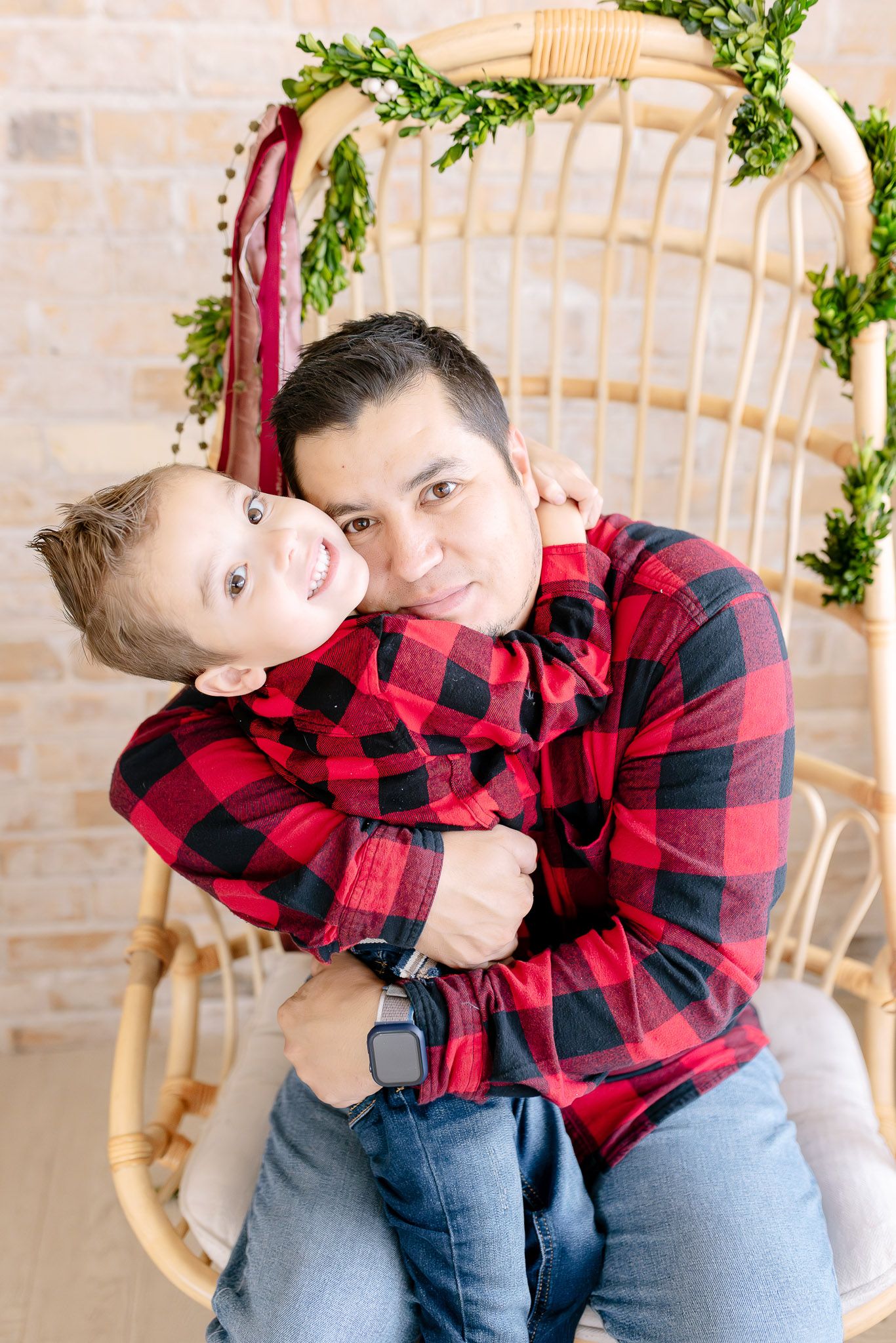 Parent and child in matching buffalo plaid shirts embrace while sitting in a rustic holiday setting.