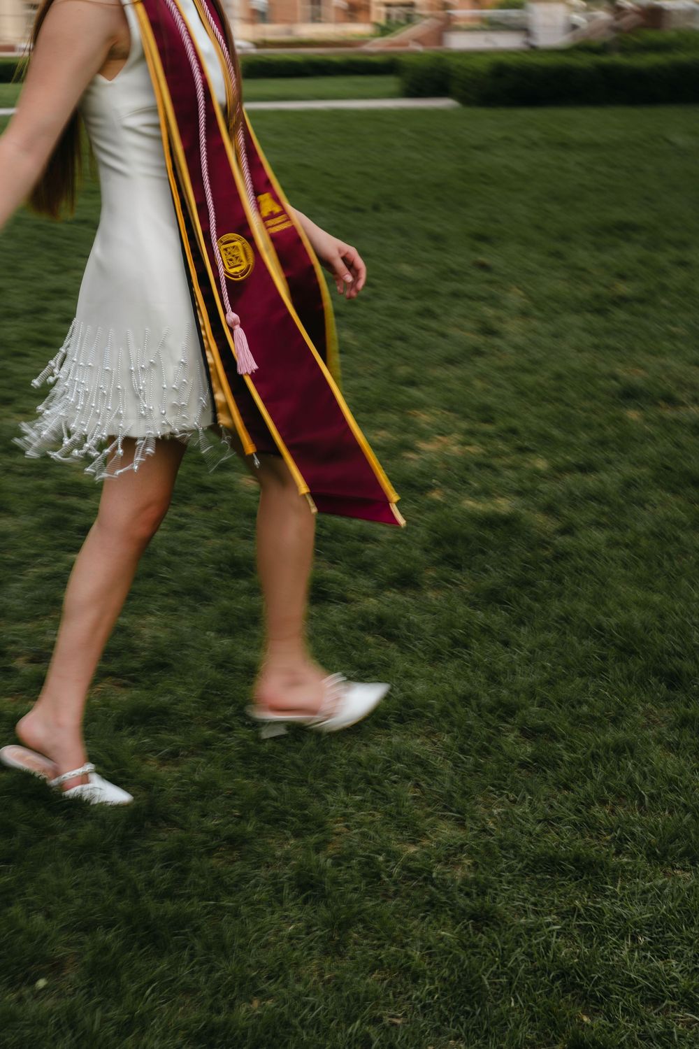 Graduates in white dresses and maroon graduation stoles walk across green grass.